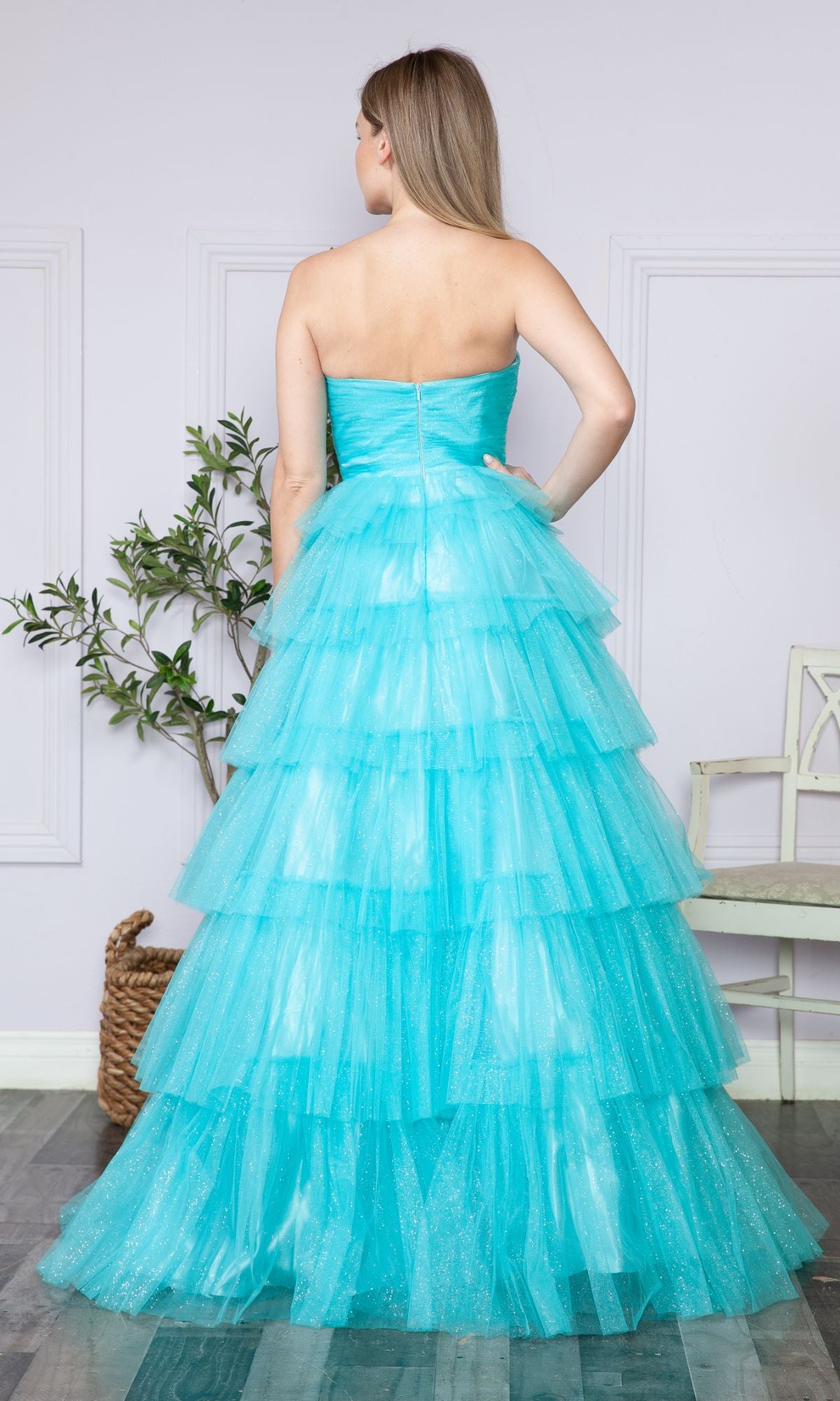 In a decorated room with a plant and chair, a woman stands with her back to the camera wearing the Strapless Long Tiered Glitter Prom Ball Gown 9386, its turquoise tulle layers sparkling in the light.